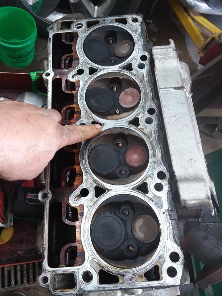 A person's finger points to a cylinder head with four combustion chambers and valves.