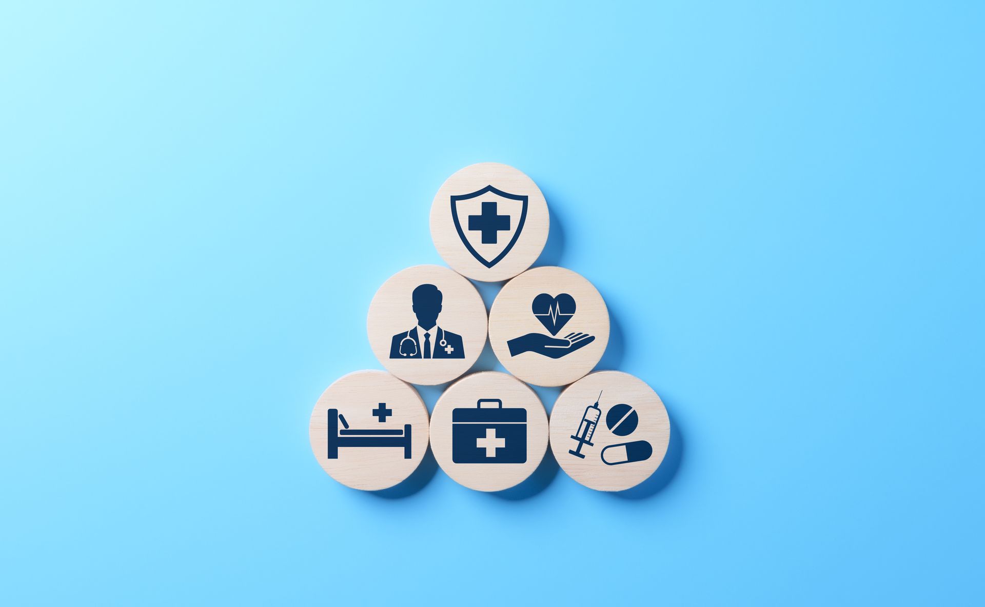 Wooden circles with health icons stacked in a pyramid: shield, doctor, heart, hospital bed, first aid kit, pills.