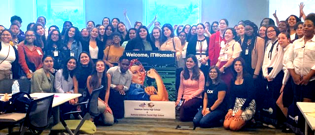 Large group posing indoors around a banner and trophy, with blue stage lighting and smiles.