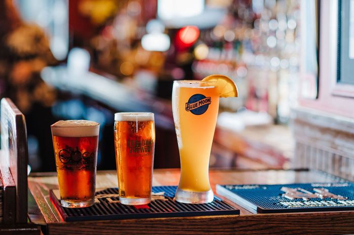 Three drinks on a bar counter, including a tall orange cocktail with a citrus garnish