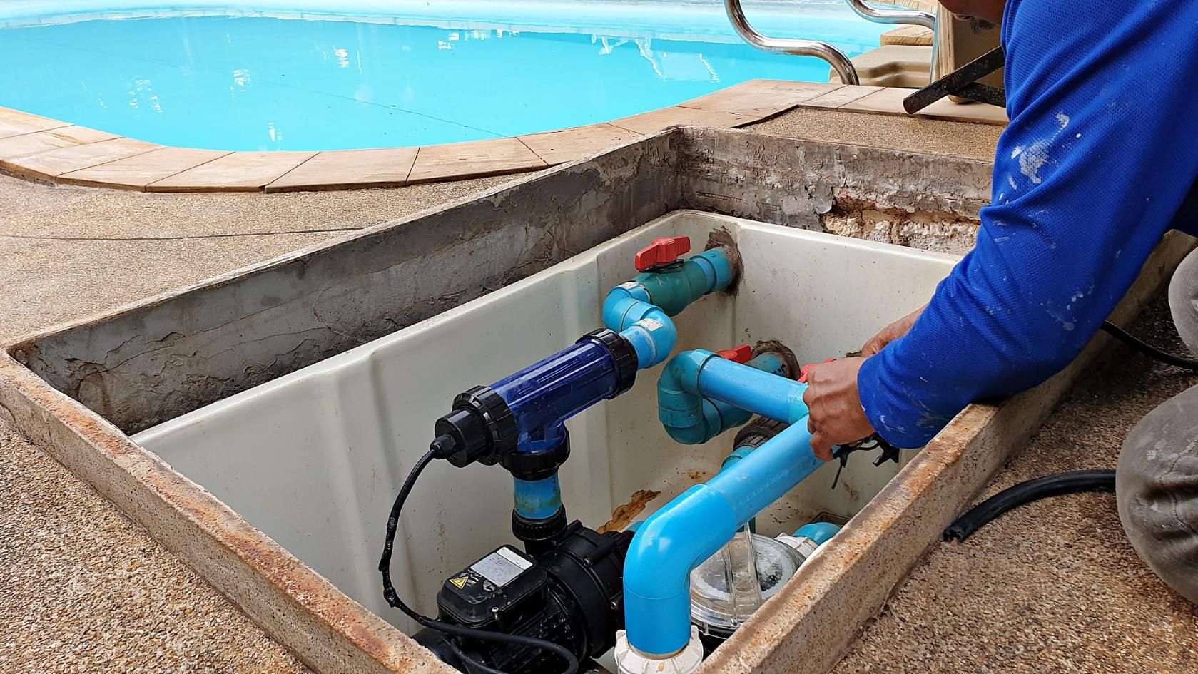 Worker installing blue pool plumbing in an open deck access hatch near a swimming pool
