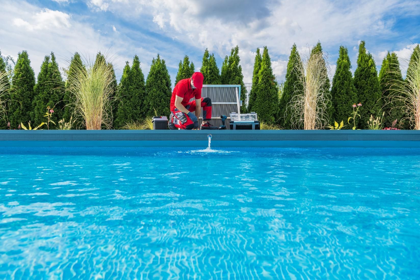 Person in red crouches by a blue pool with trees and sky in the background