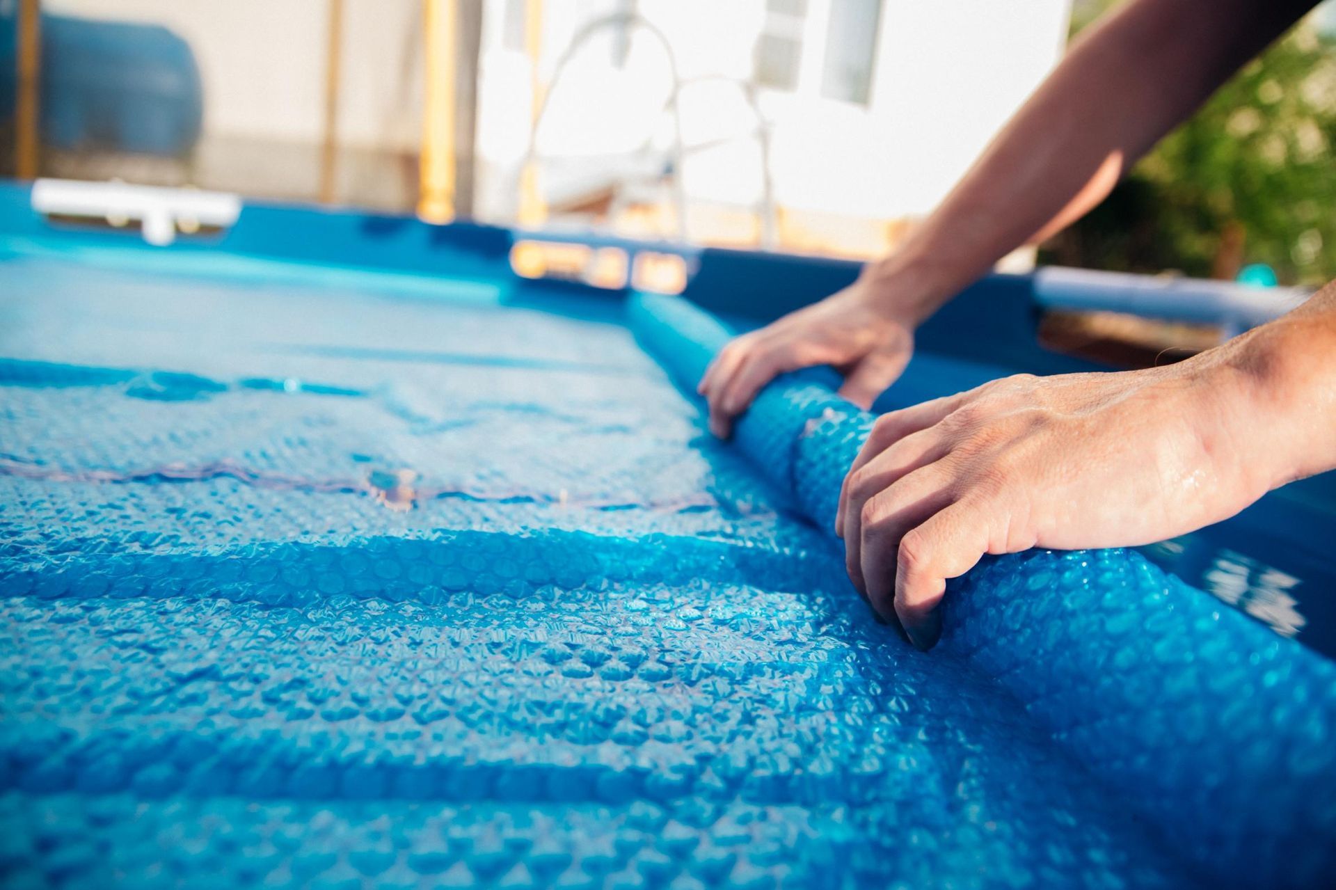 Hands rolling up a blue pool cover beside a swimming pool.