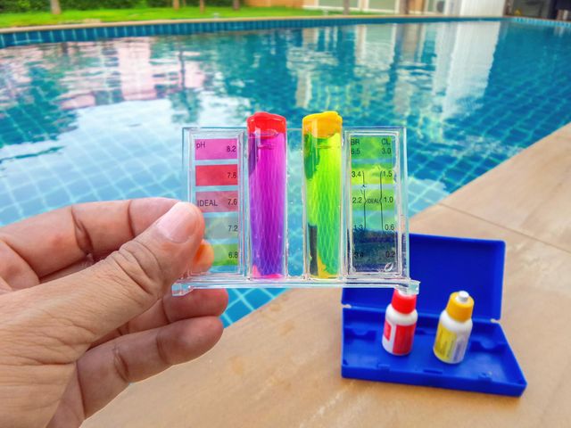 Hand holding colorful test tubes by a swimming pool, with small bottles on a blue stand