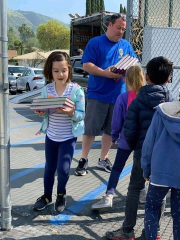 A group of children are standing around a man holding a stack of books.