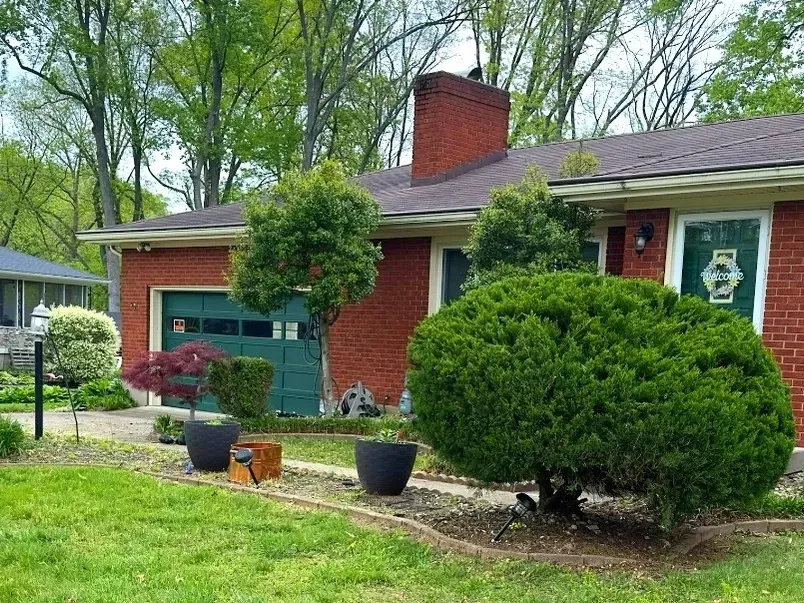 A brick house with a green garage door is surrounded by trees and bushes.