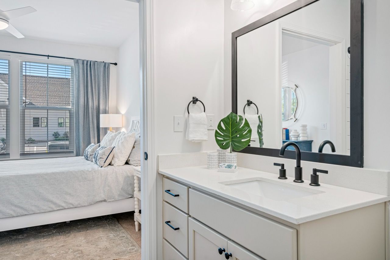Bright bedroom with a large window, white bedding, and a nearby bathroom vanity with a mirror.