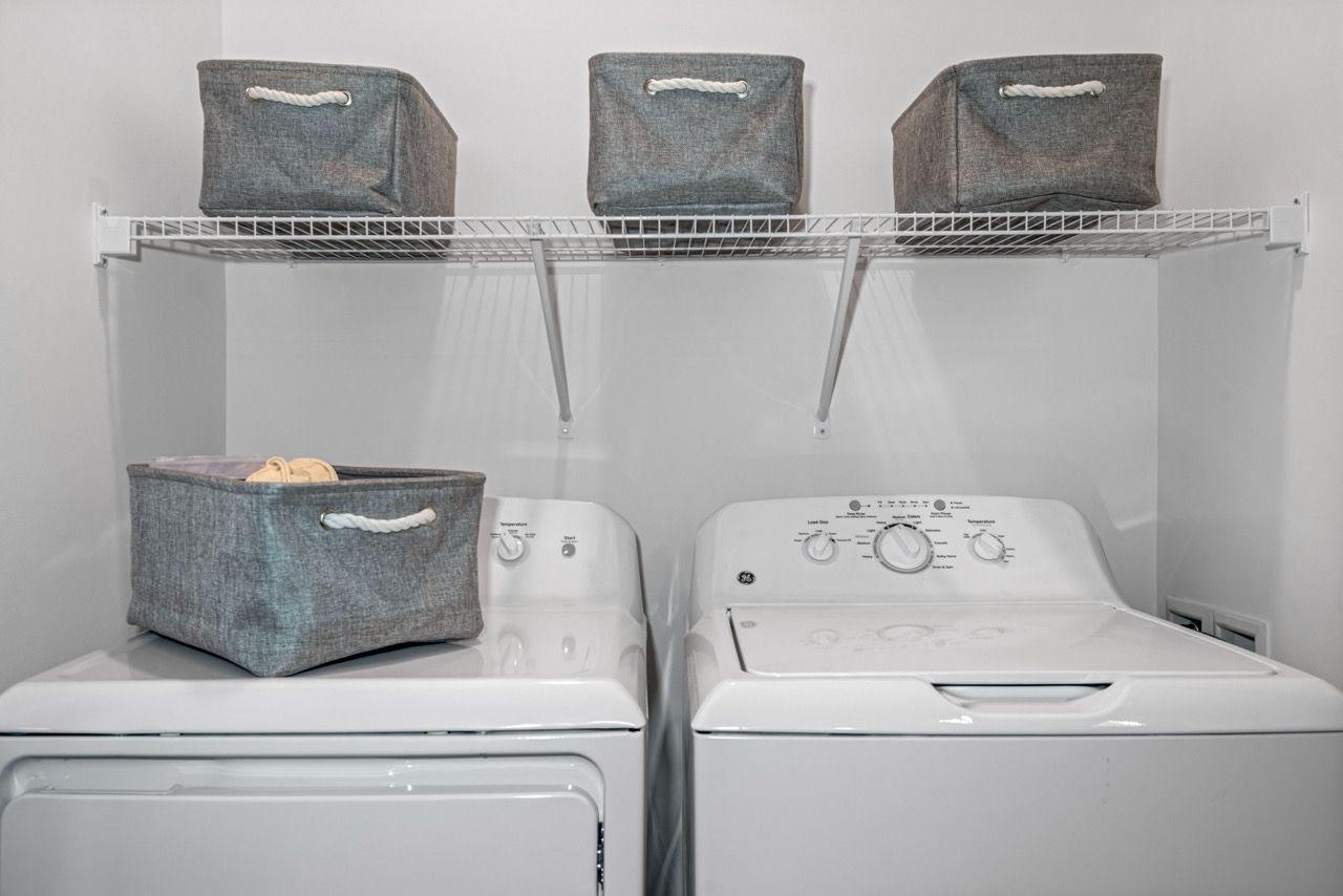 Laundry area with a washer and dryer and wire shelf holding gray fabric storage bins.