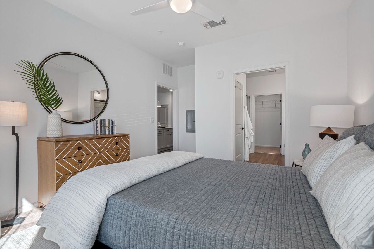 Bedroom in a modern apartment with gray bedding, a round mirror above a geometric dresser, and a closet doorway.