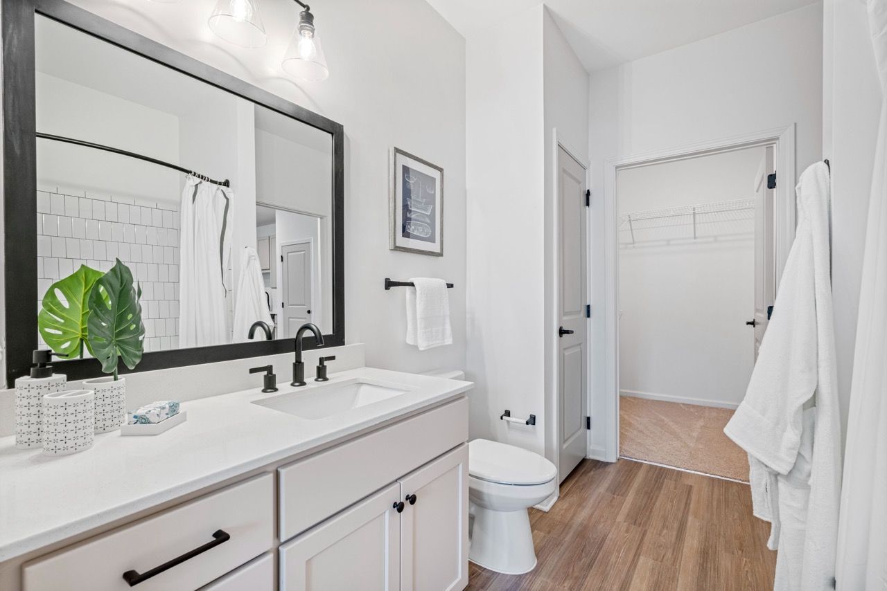 Bright white bathroom with a white vanity, black faucet, large mirror, and an open walk-in closet visible behind.