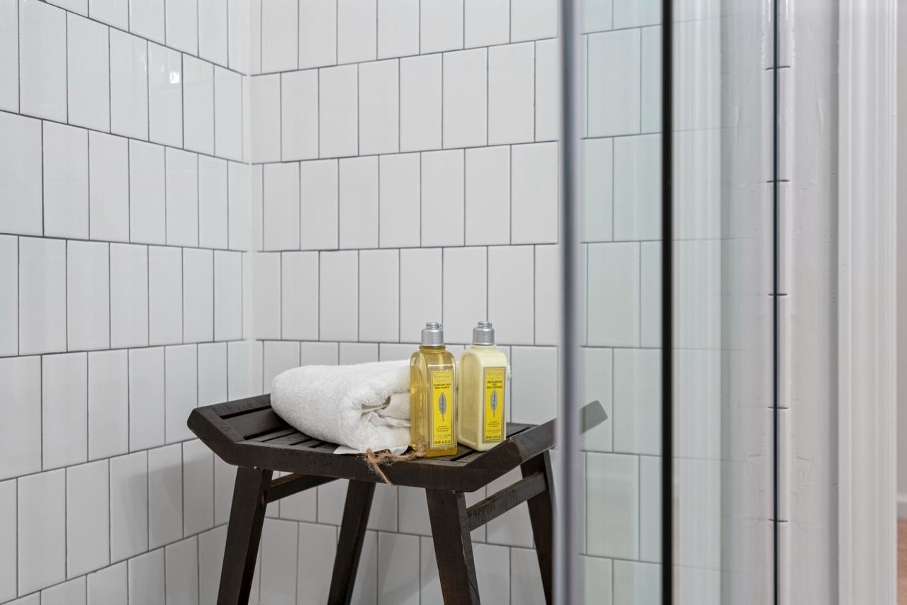 White tiled bathroom with a dark wooden stool, folded towel, and two bottles beside a glass shower door.