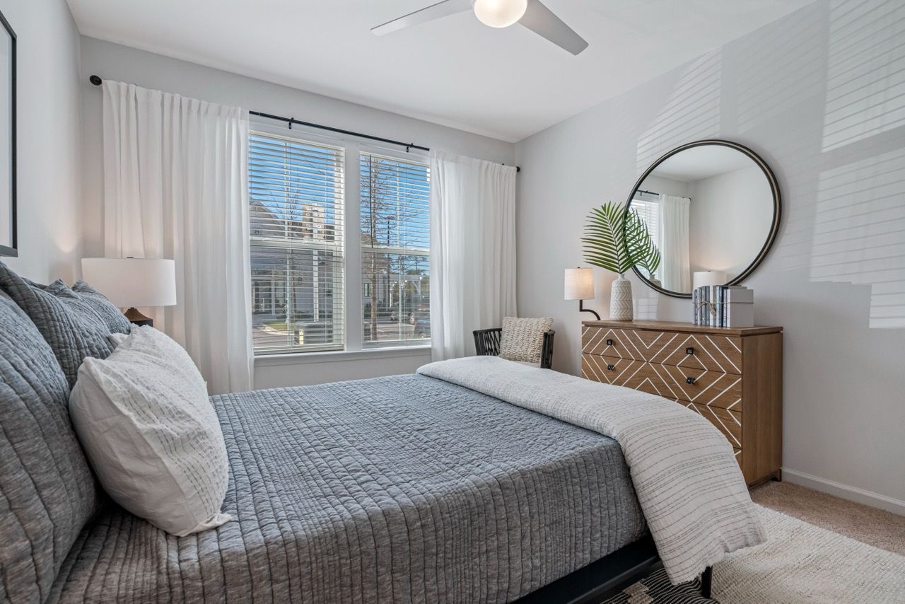 Bedroom with a gray quilted bed, white pillows, dresser with round mirror, and a large window with white curtains.