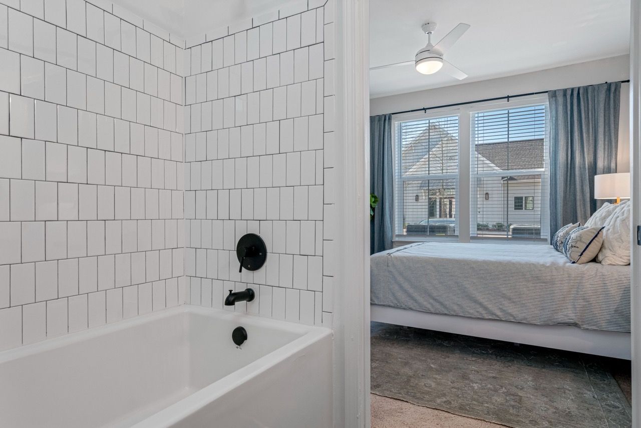White tiled bathroom tub area with black fixtures, opening to a bedroom with a window and blue curtains.