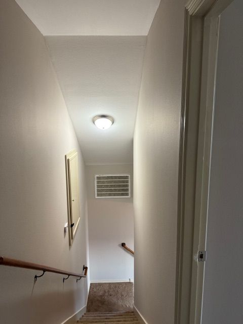 A narrow indoor staircase with light-colored walls, a wooden handrail, a ceiling light, and a rectangular wall vent.