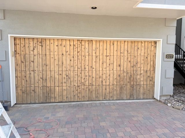 A horizontal, wooden-slatted garage door with a light brown, natural wood finish in a gray stucco residential setting.