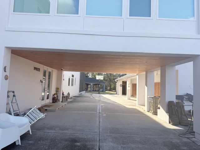 A concrete driveway passes under a white modern building structure with a wood-paneled ceiling and adjacent outdoor space.