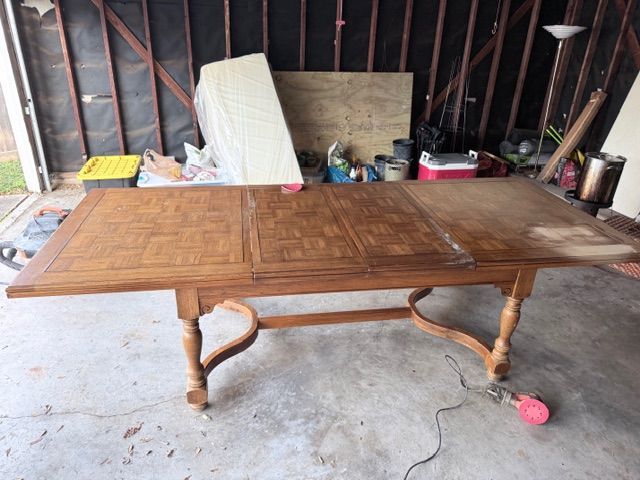 An unfinished wooden dining table with a parquet top stands in a garage, next to an electric sander on the concrete floor.