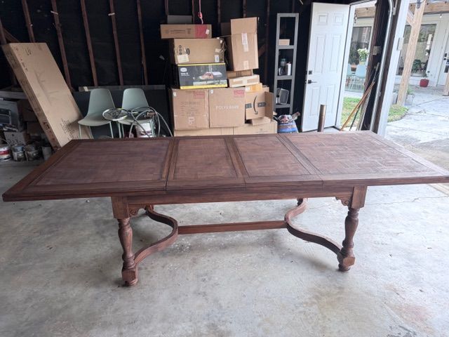 A wooden dining table with a center extension leaf and carved legs, positioned inside a garage with boxes in the background.