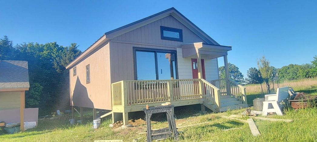 Small blue cabin with porch in a sunny grassy yard, with trees and a neighboring building nearby