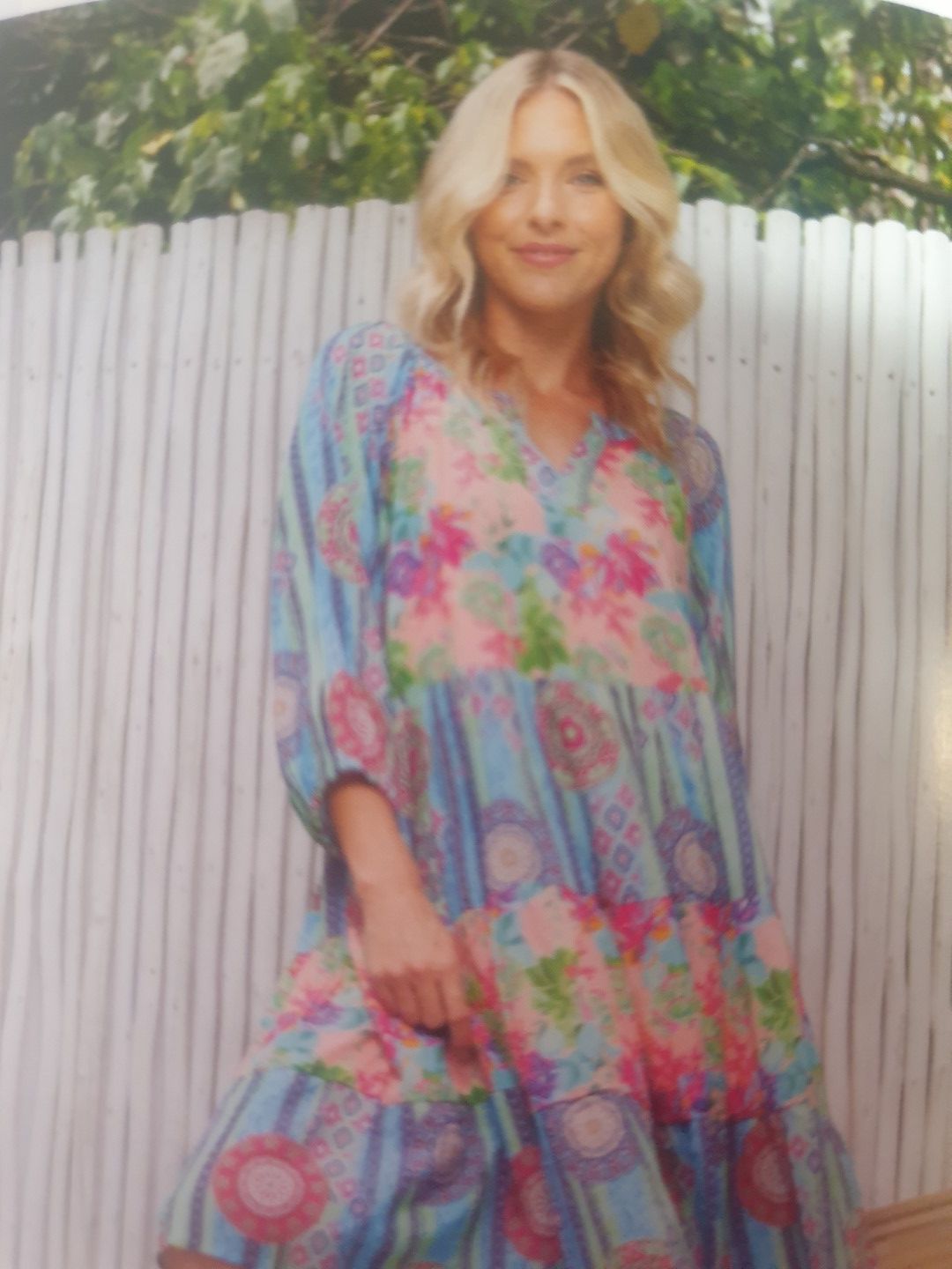 Blonde woman in floral dress smiles, standing in front of a white slatted fence.