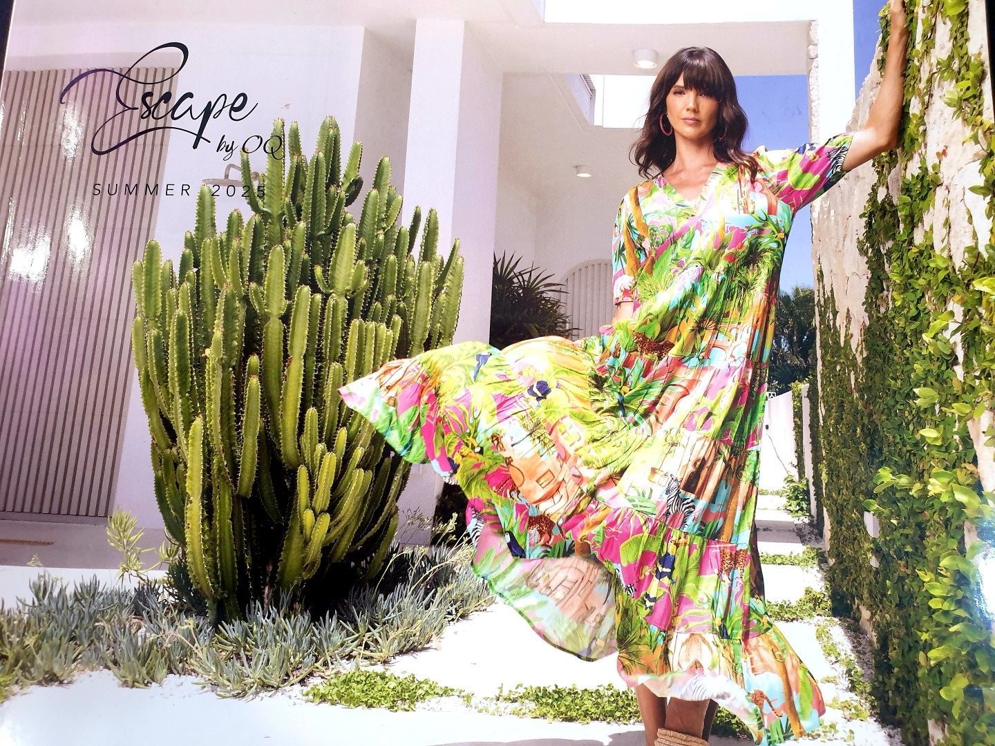 Woman in colorful dress leaning on a wall by greenery; outside a white building.