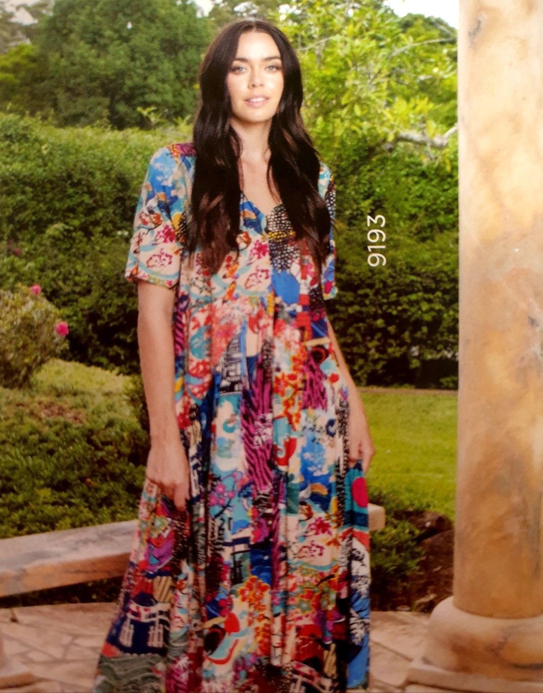 Woman in a colorful printed dress stands outdoors, smiling.