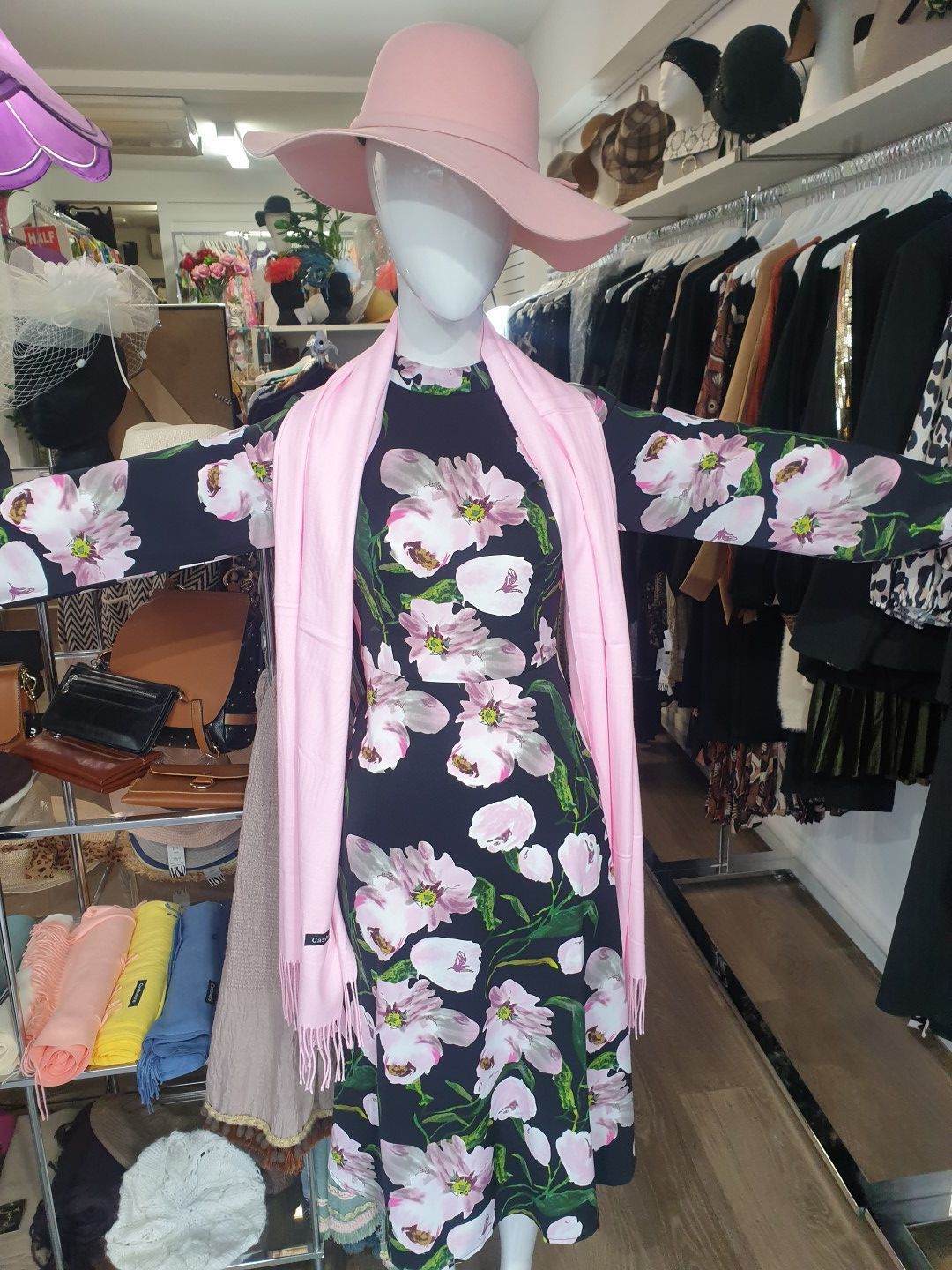 Mannequin in floral dress, pink scarf, and hat, in a clothing store.