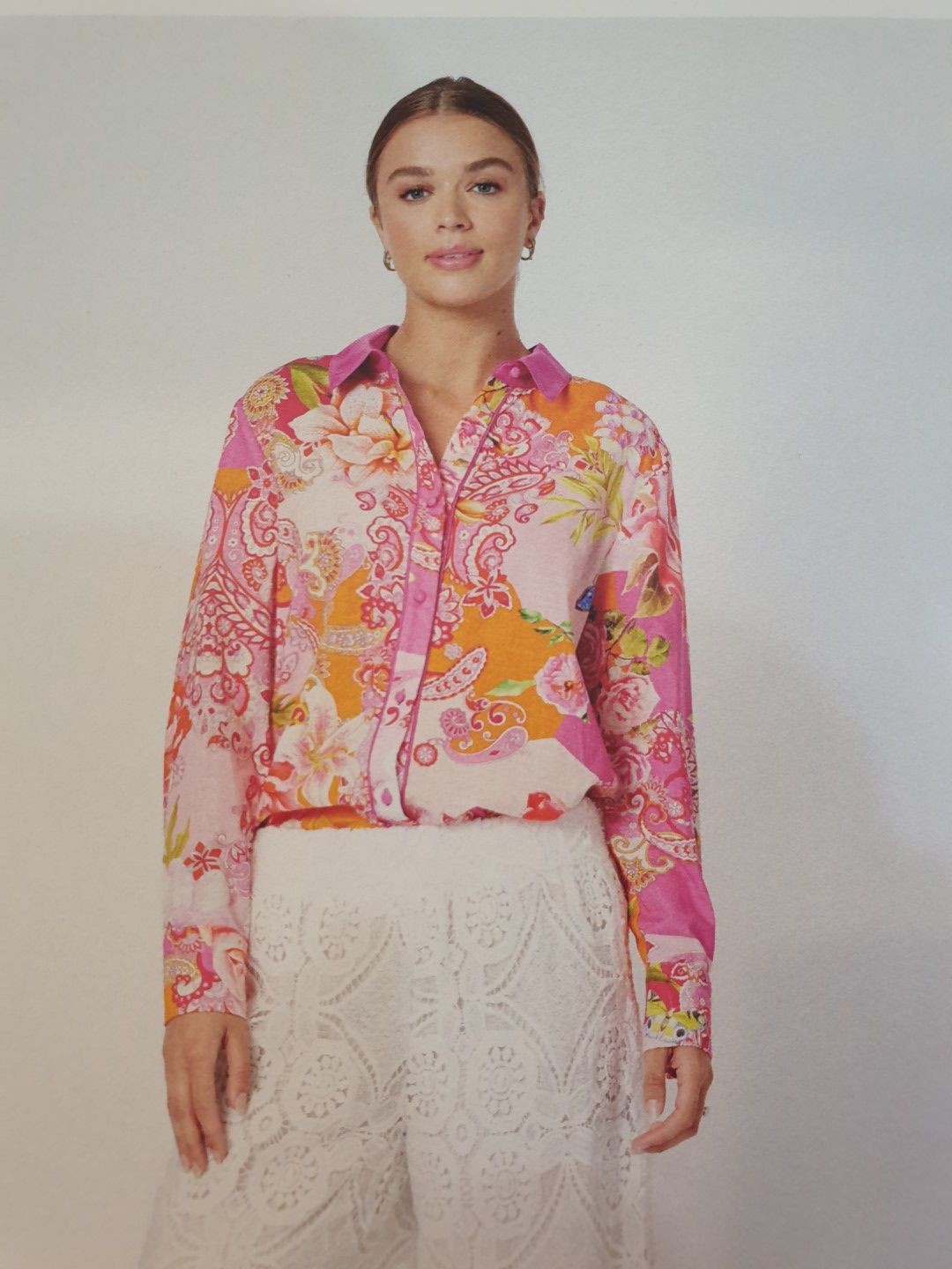 Woman wearing a colorful floral shirt and white lace pants, smiling. Indoor setting, neutral background.