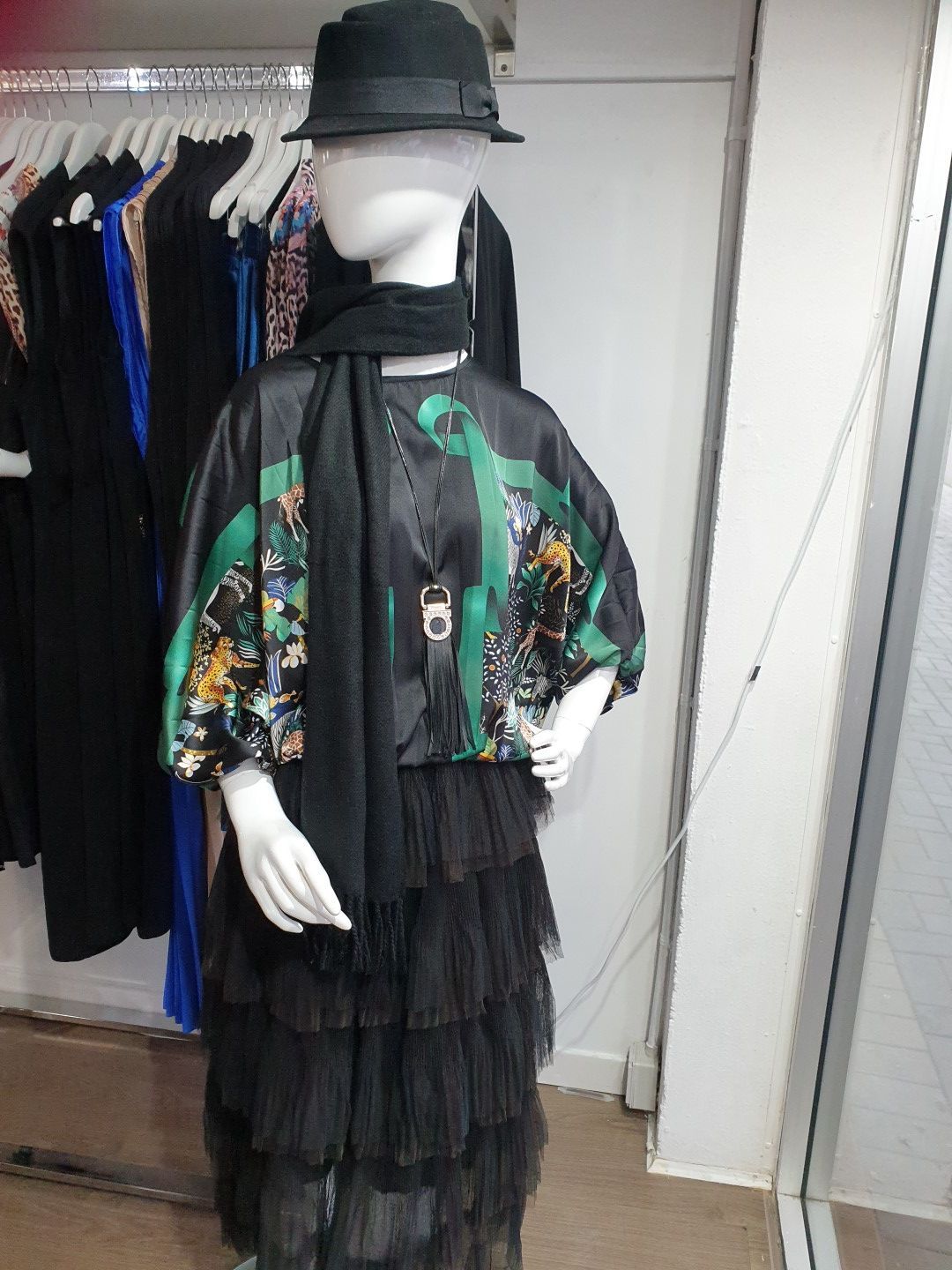 Mannequin in a black outfit with a tiered skirt, green floral top, scarf, hat, inside a clothing store.