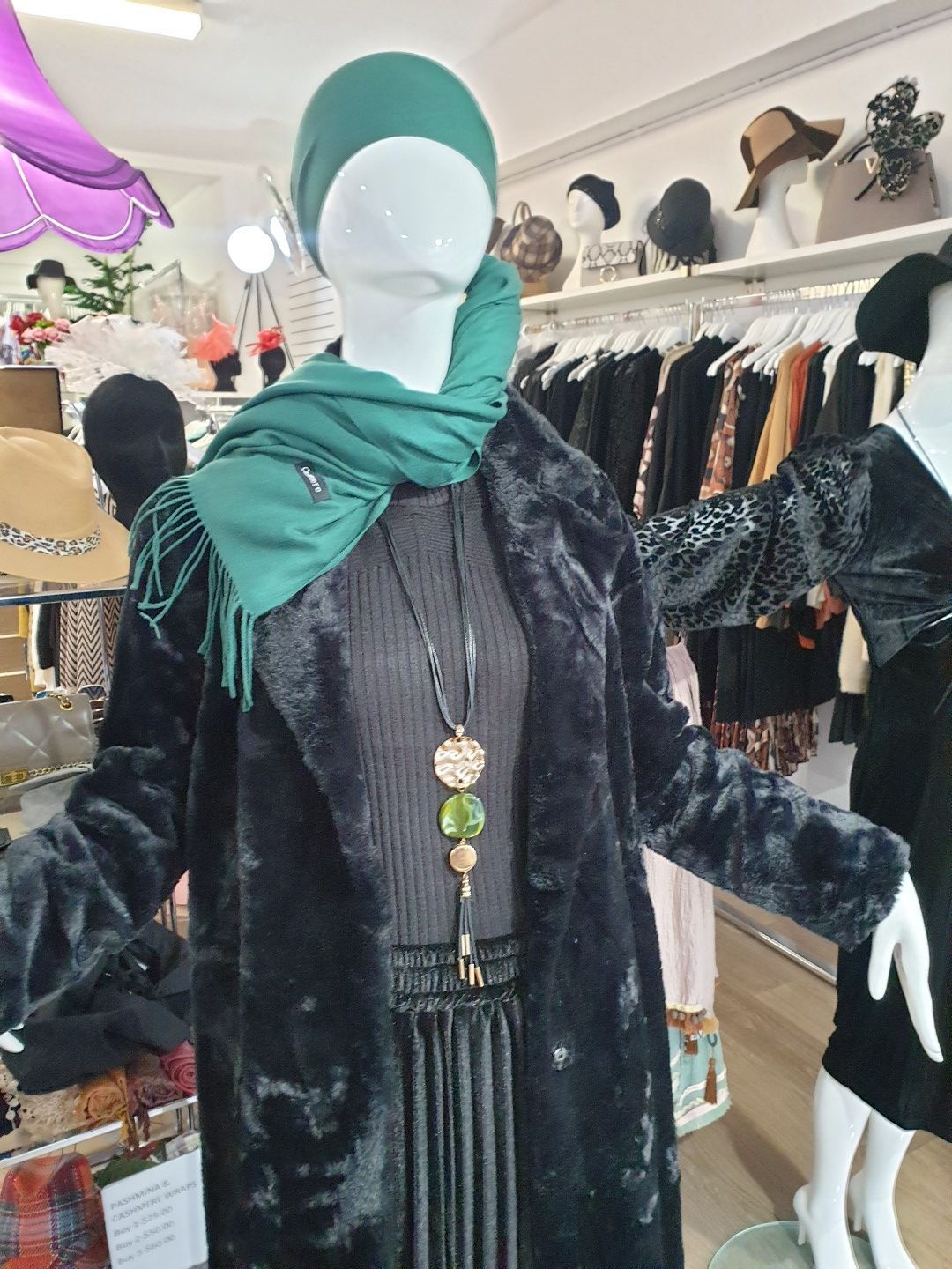 Mannequin in black faux fur coat, green scarf and hat, black dress, store interior.