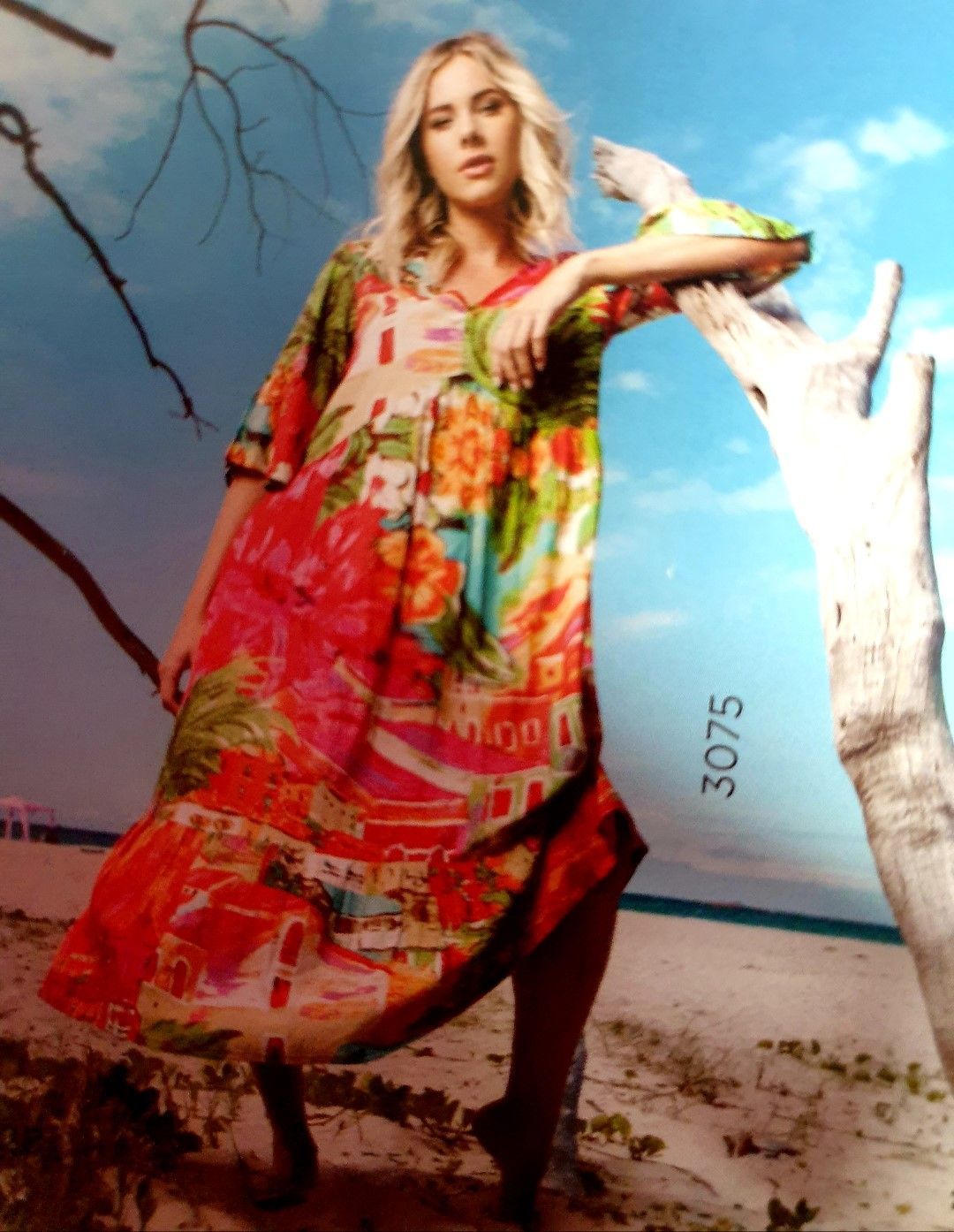 Woman in colorful dress leans on a tree, on a beach, with a blue sky background.
