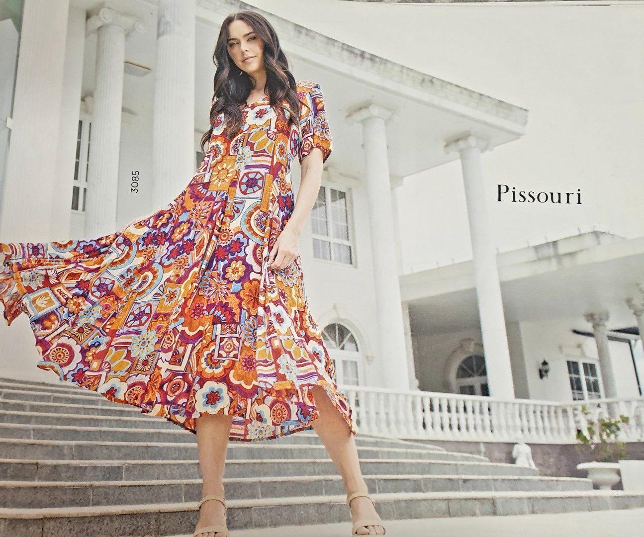 Woman in a patterned orange and purple dress poses on steps in front of a white building.