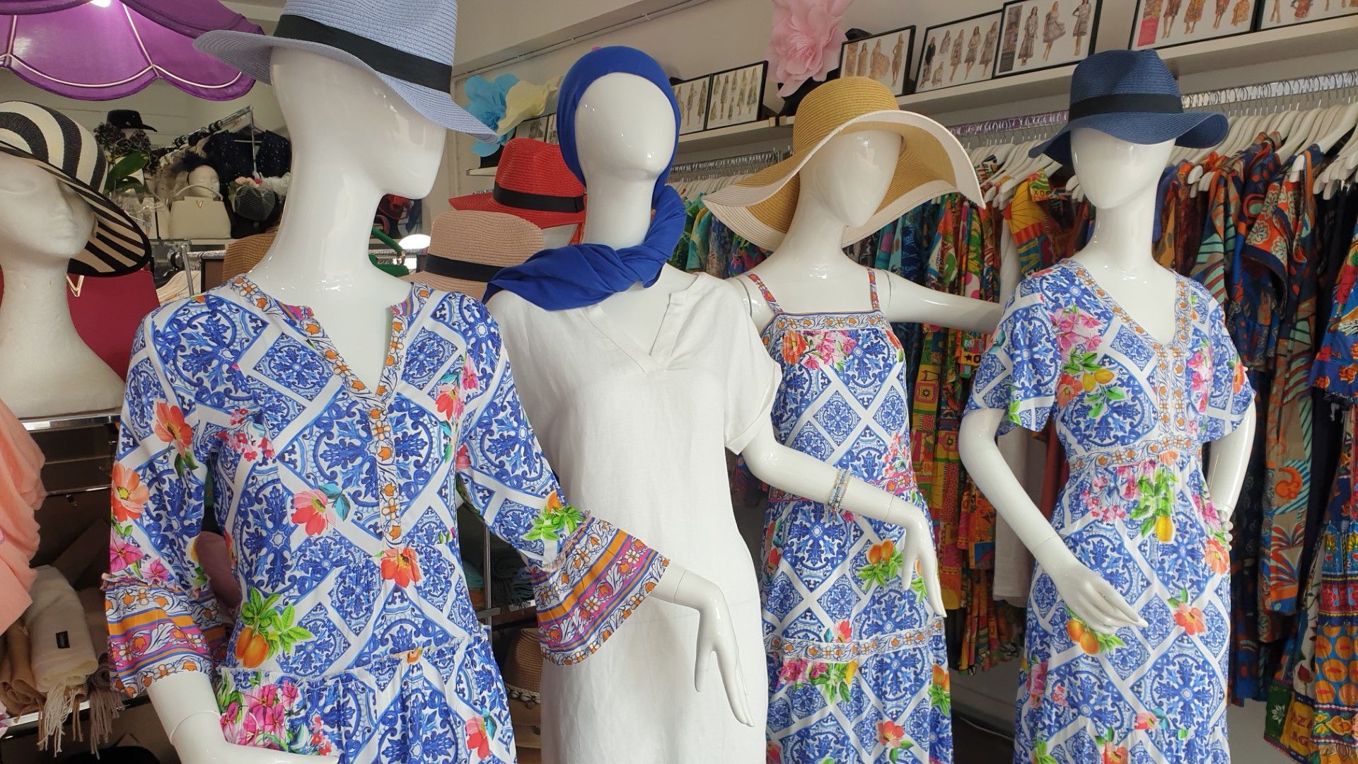 Mannequins in a clothing store display summer dresses and hats; various colors, background racks of clothing.