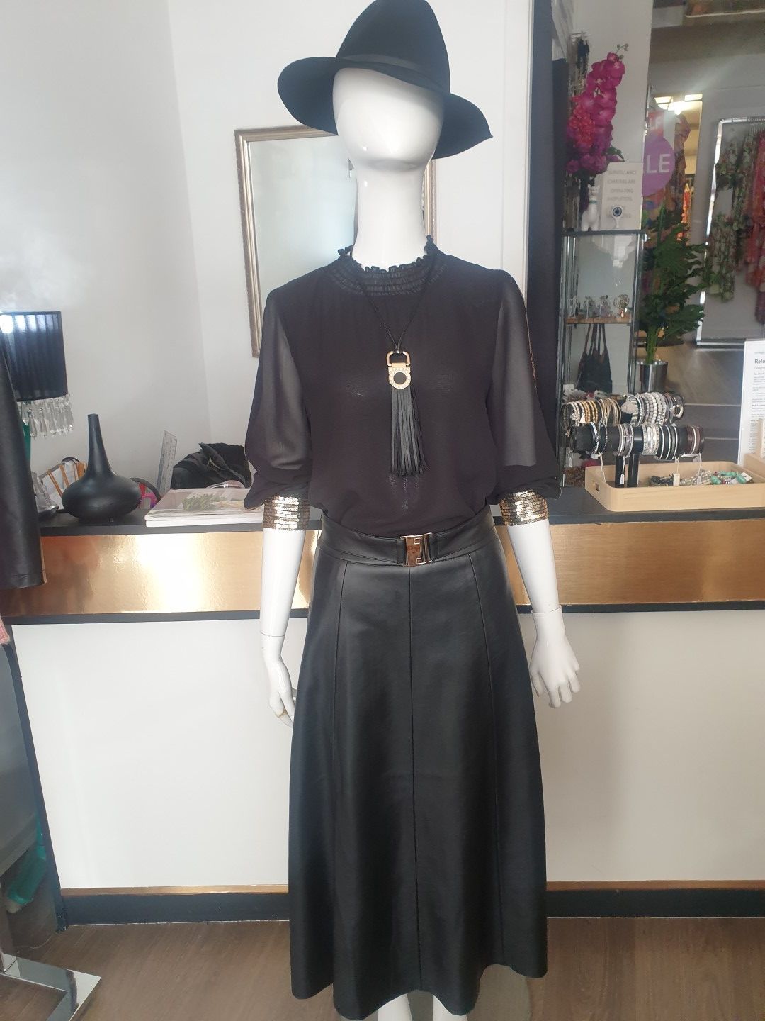 Mannequin in black outfit: hat, sheer-sleeved top with tassel, leather skirt, gold accents. Store interior.