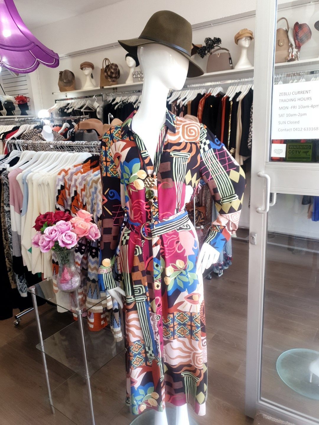 Mannequin in patterned dress and hat, inside clothing store. Racks of clothing and accessories in the background.