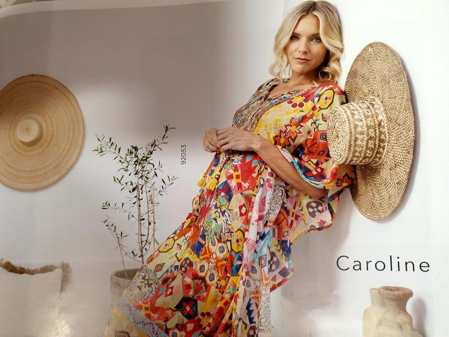Woman in colorful dress leans against a wall; straw hat hangs next to her.