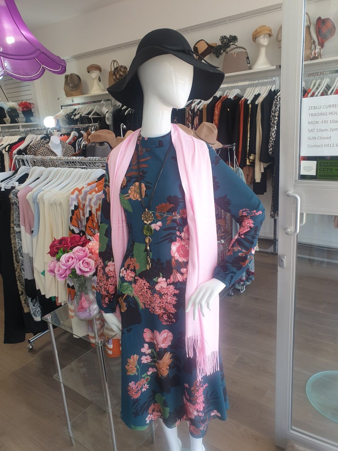 Mannequin in floral dress, black hat, pink scarf, inside clothing store, various clothing racks.