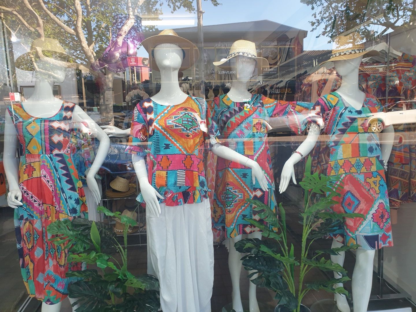 Four mannequins in colorful patterned dresses and straw hats in a shop window display.