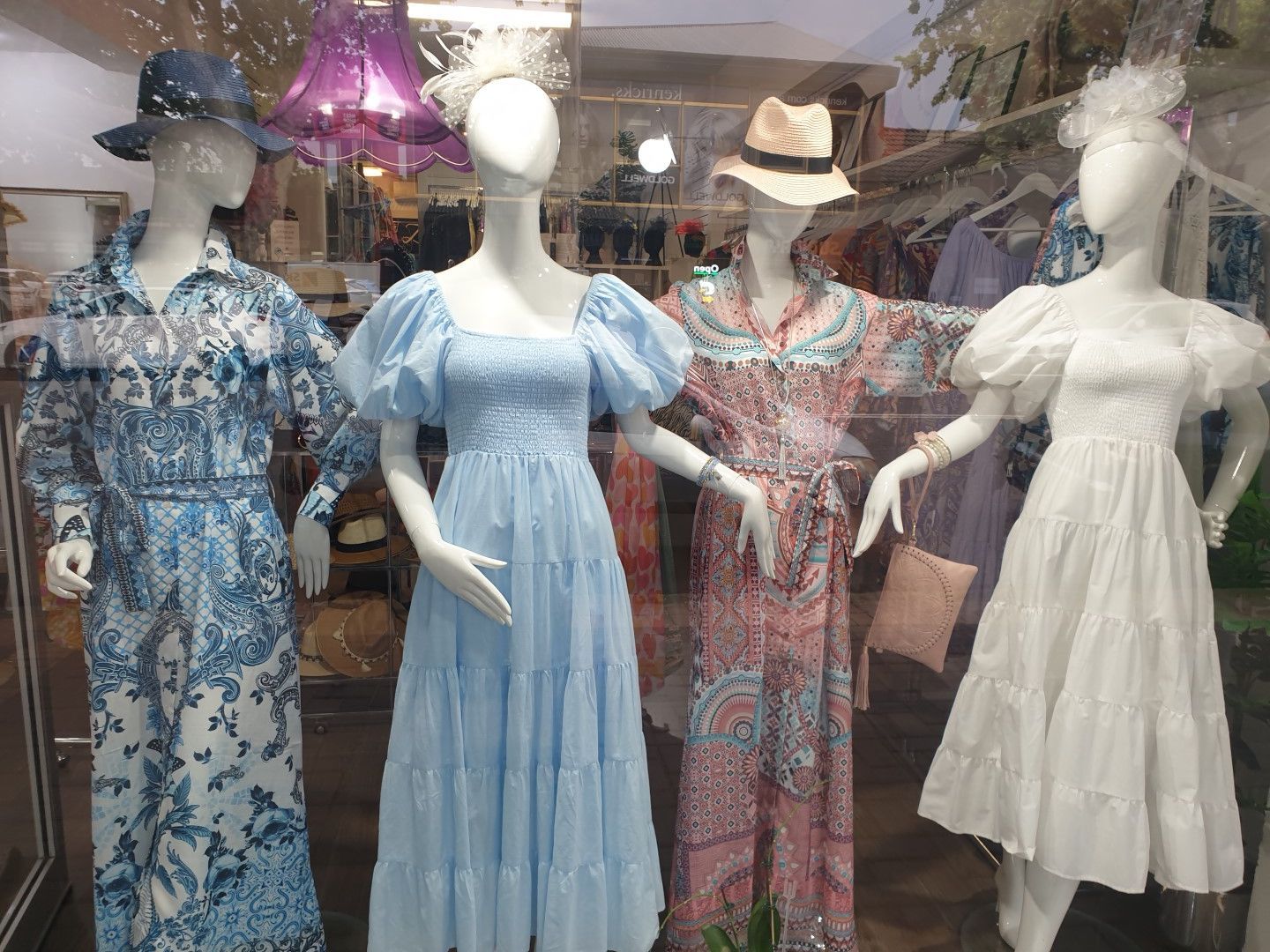 Four mannequins in a shop window, each wearing a dress and hat, in shades of blue, pink, and white.