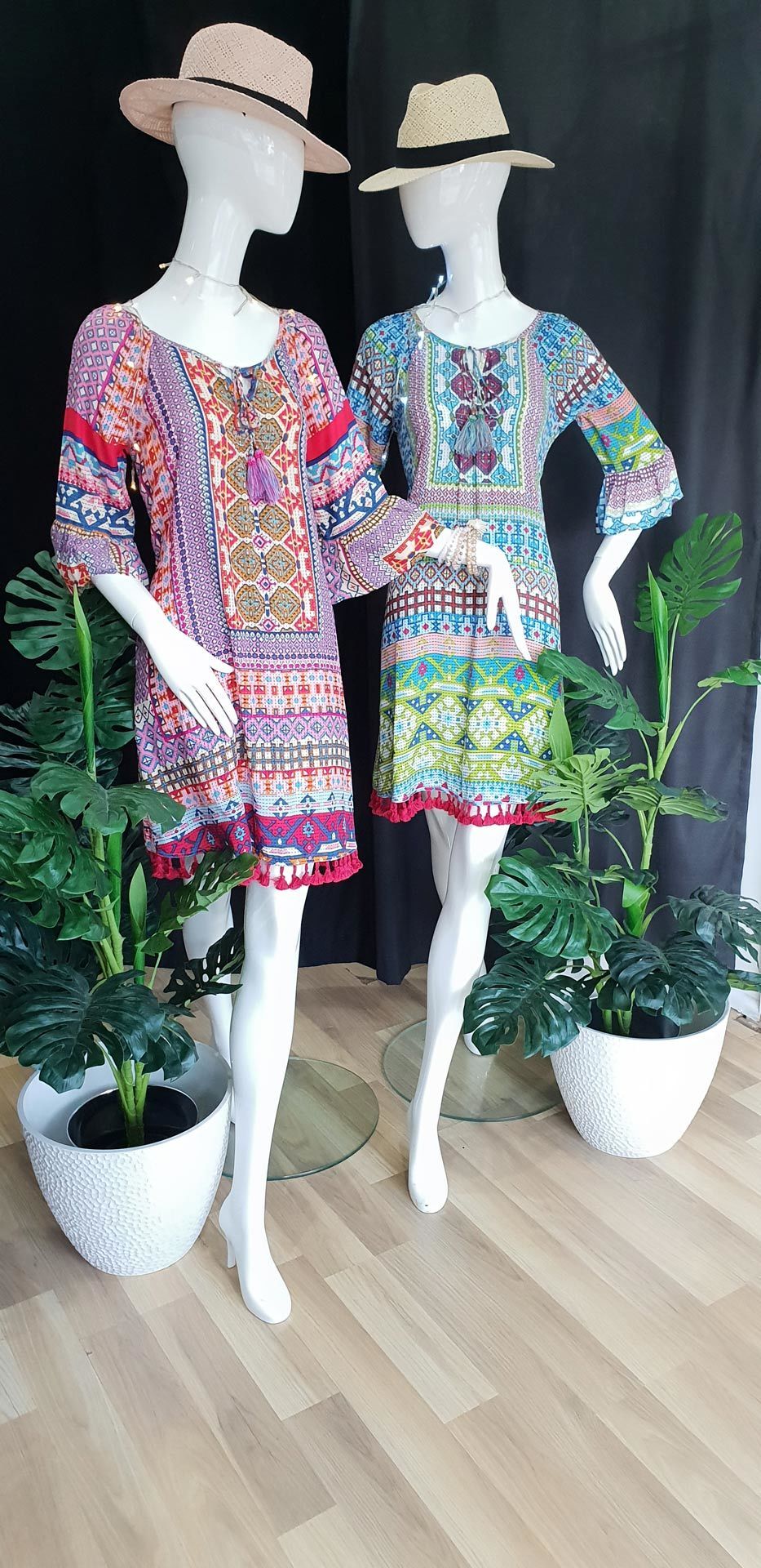 Two mannequins wearing colorful dresses and hats, flanked by potted plants, indoors.