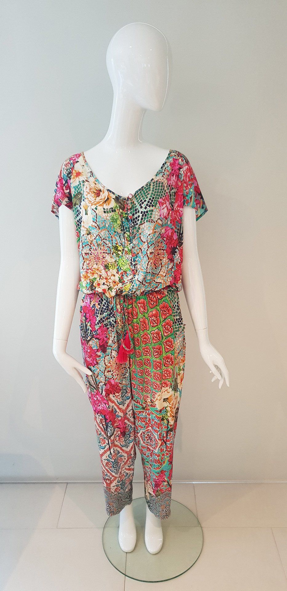 Mannequin wearing a floral print jumpsuit; standing against a pale blue wall, on a glass stand.