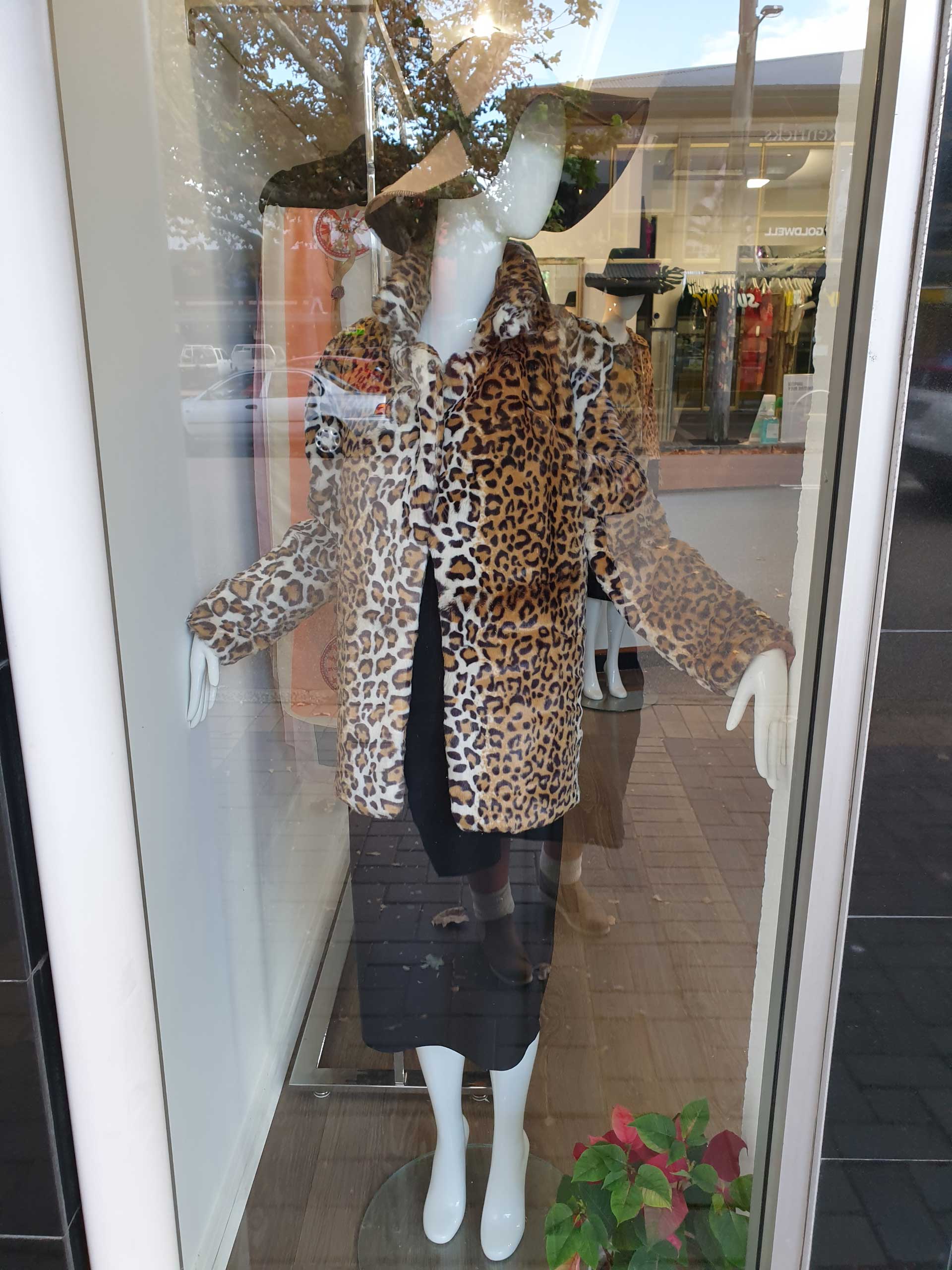 Mannequin in a shop window wearing a leopard print jacket, black dress, and skirt. White feet.