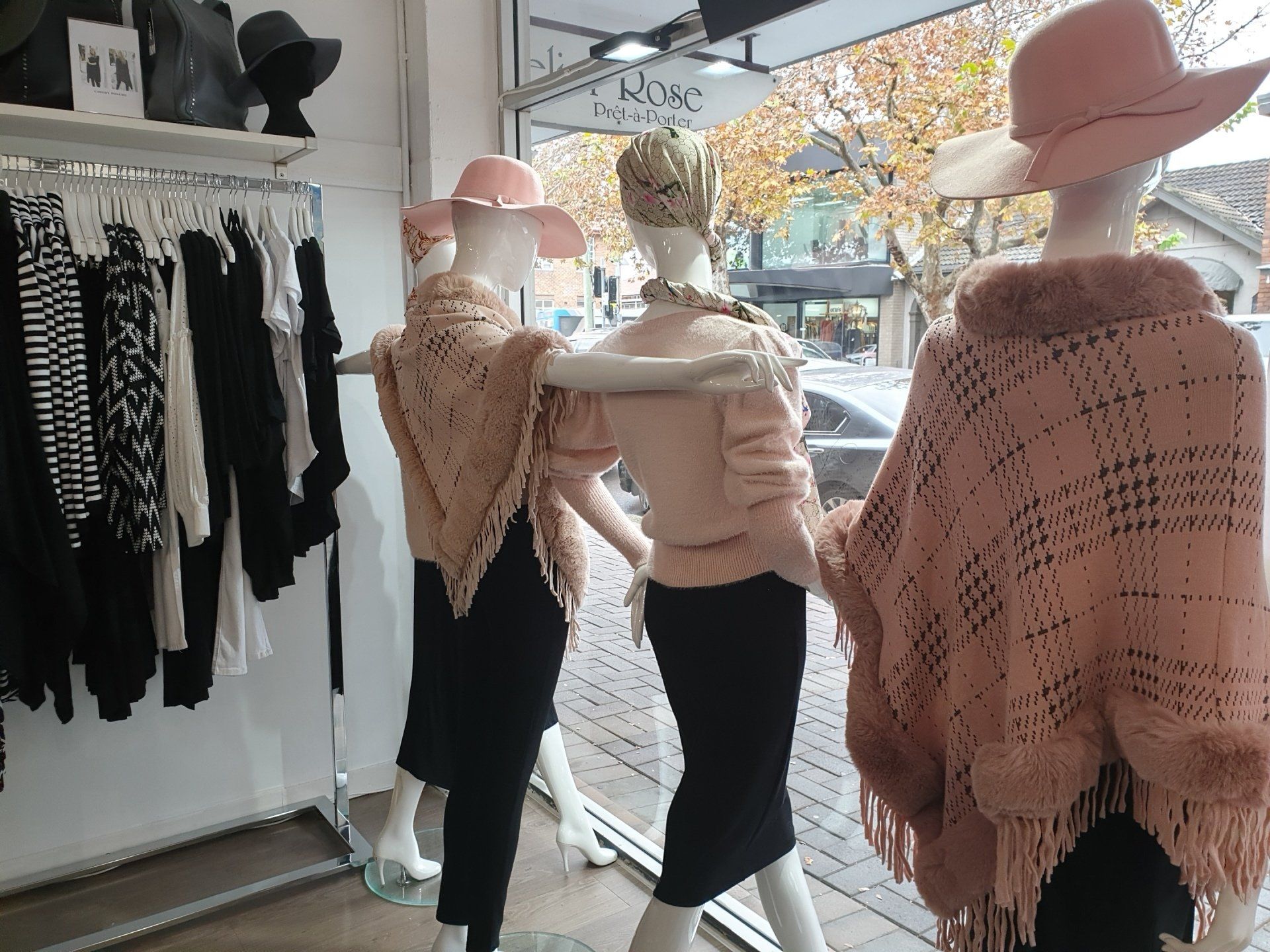 Clothing store window display with mannequins wearing pink hats and shawls. Racks of black and white clothes.