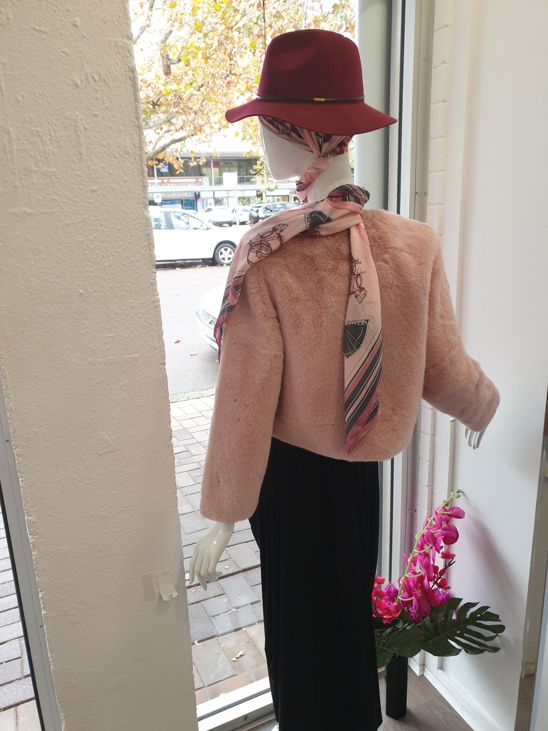 Mannequin wearing a pink sweater, black skirt, maroon hat, scarf. View of street outside the window.
