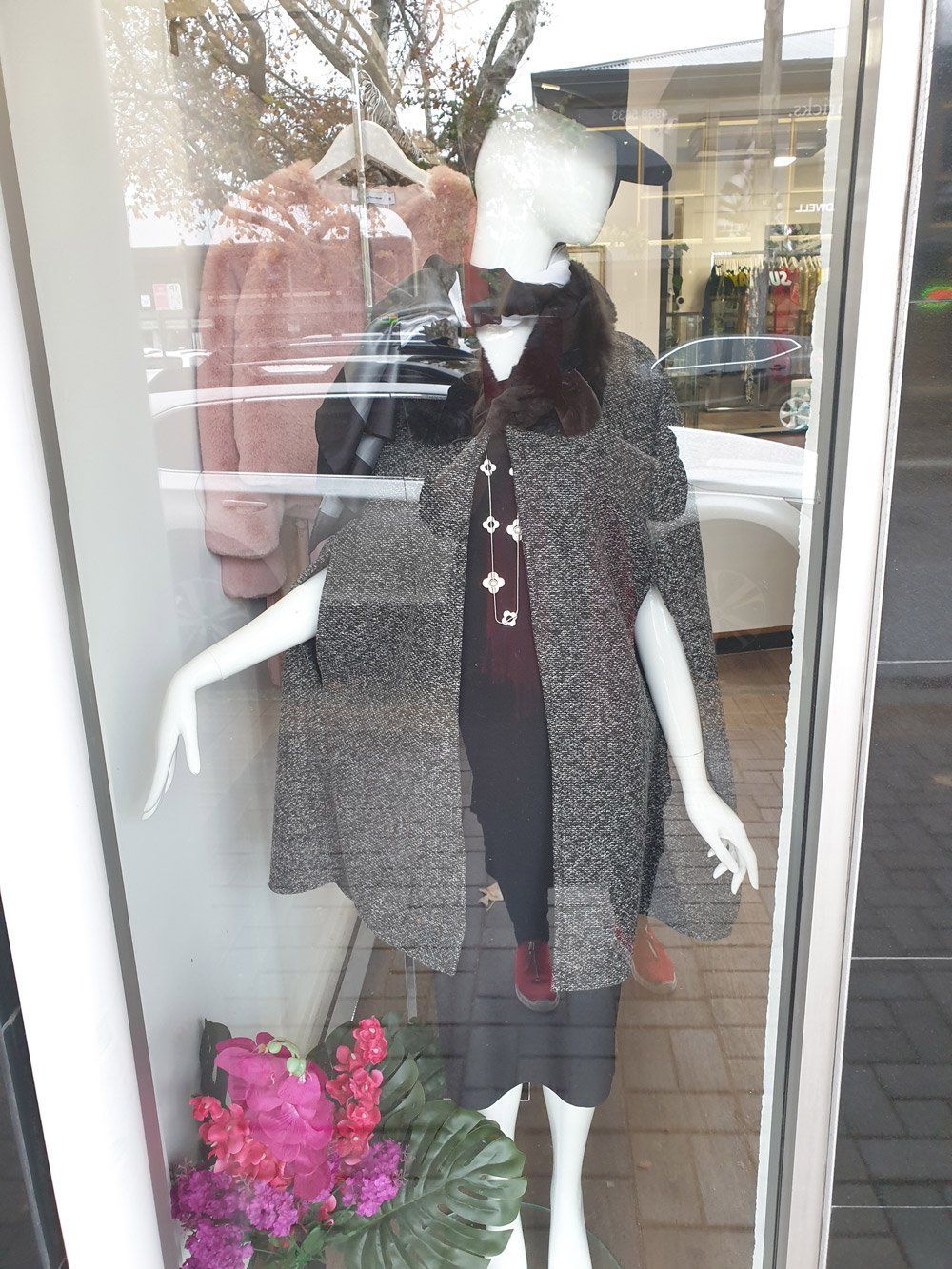 Mannequin in store window wearing cape coat with fur collar; floral display.