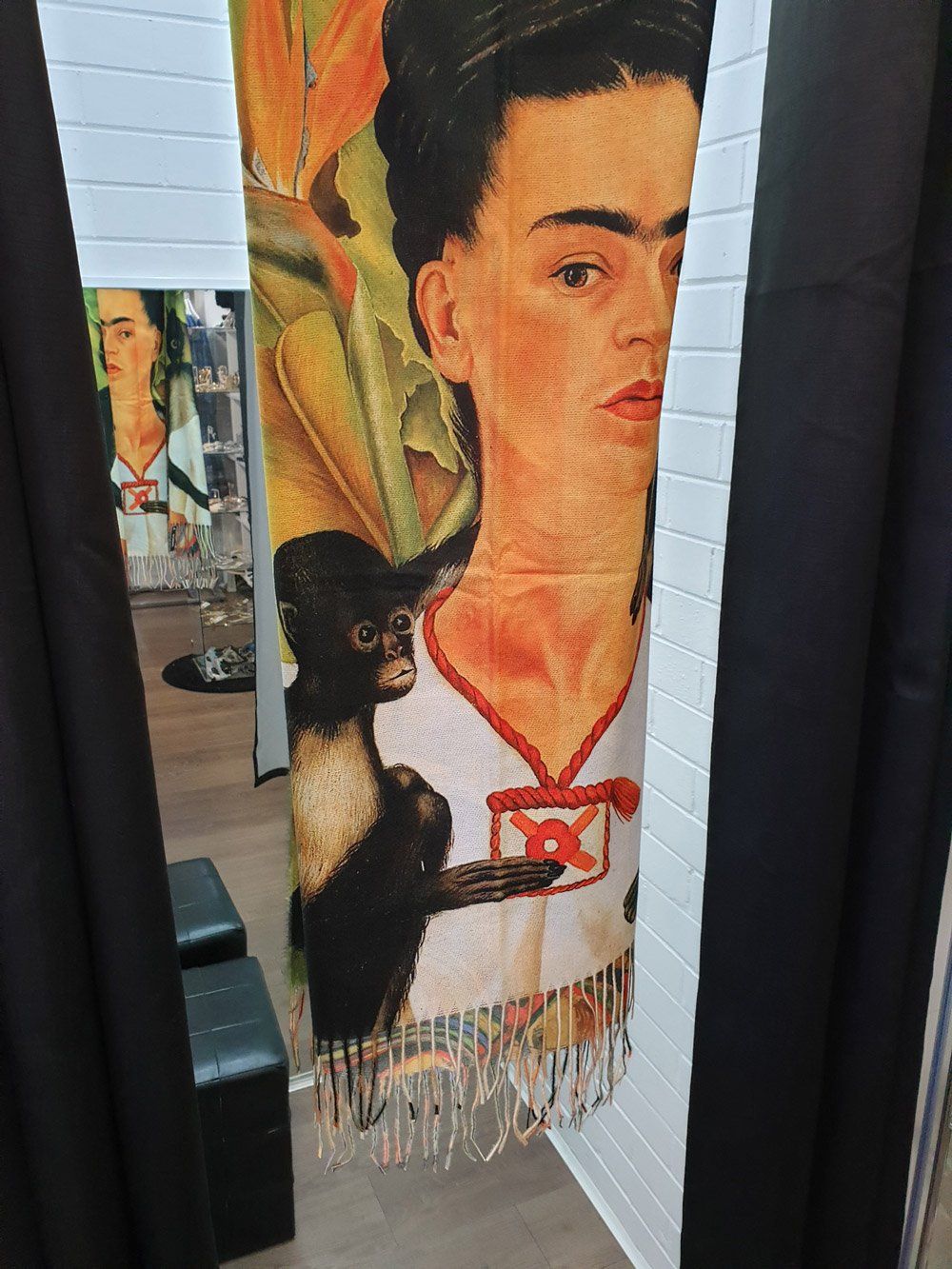 Scarf with Frida Kahlo portrait and monkey, fringed edges. Seen through black curtains, white wall background.