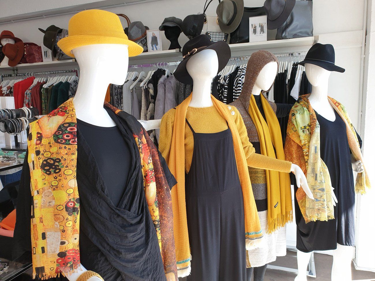 Four mannequins in a store window display, wearing hats and colorful scarves and clothing, clothes rack in background.