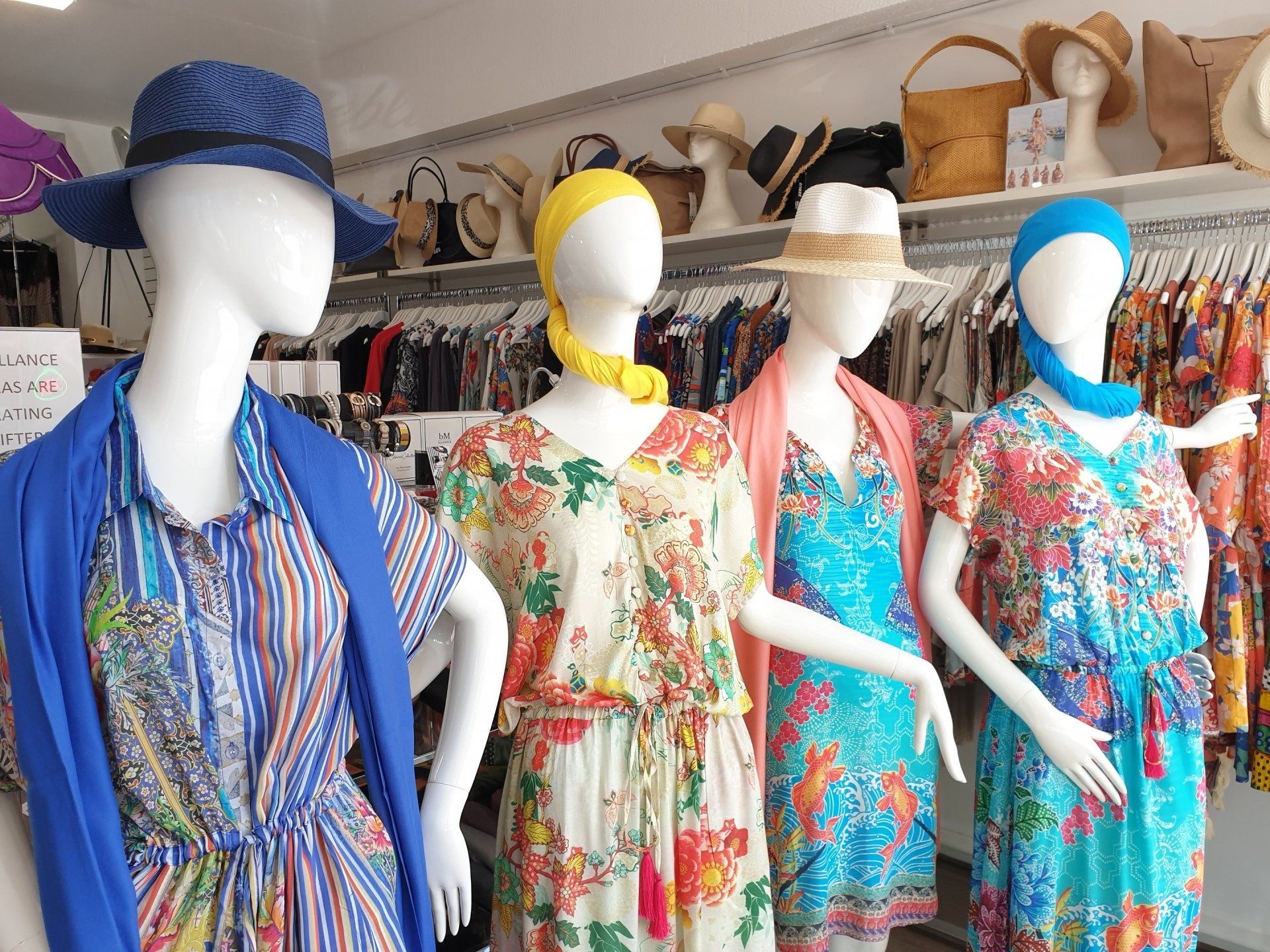 Mannequins in a clothing store, wearing colorful dresses and hats. White walls and shelves with more hats in the background.