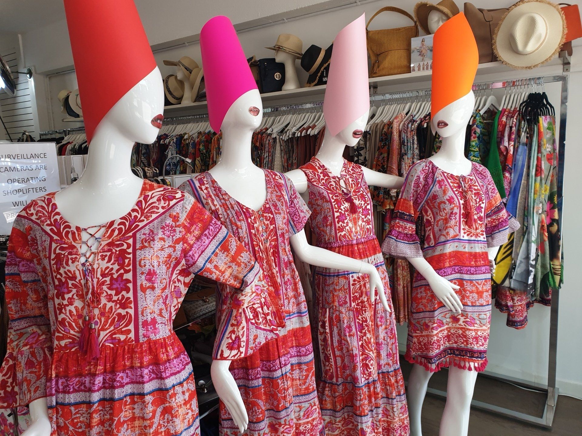 Four mannequins in red floral dresses and colorful cone-shaped hats, in a clothing store.