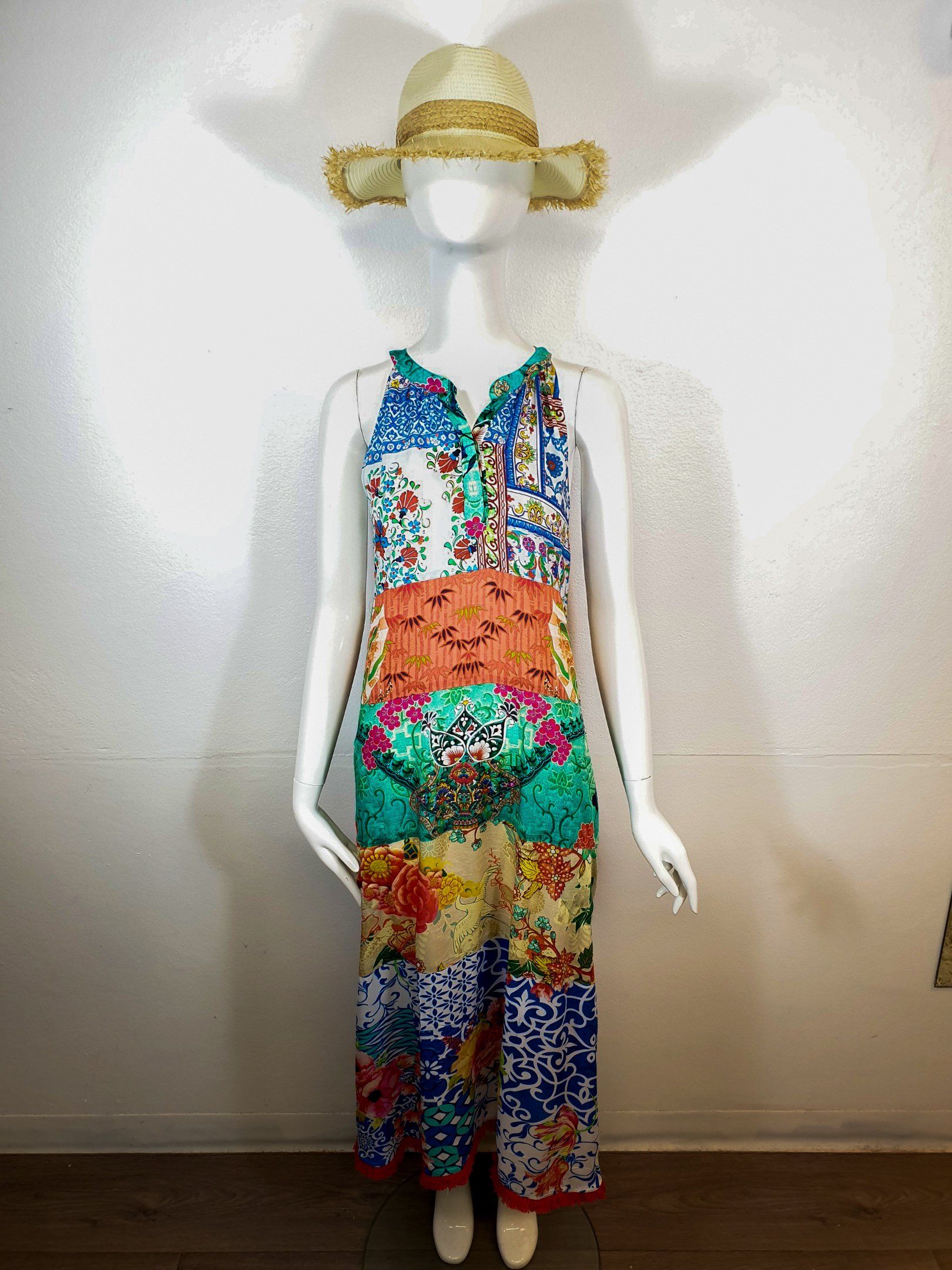 Mannequin wearing a colorful patterned maxi dress and straw hat. Standing in front of a white wall.